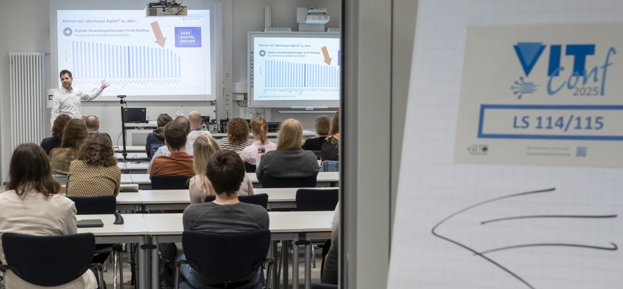 Prof. Dr. Sebastian Halsbenning hält die KeyNote vor dem Auditorium von Ehemaligen, Studierenden und Lehrenden. Rechts neben der Tür, durch die das Foto aufgenommen wurde, ist ein Hinweisschild zur VITConf zu sehen.